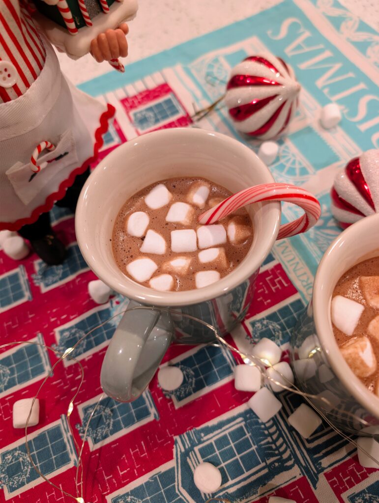 A white ceramic mug filled with hot cocoa and marshmallows sitting next to Christmas ornaments and fairy lights.