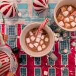 Overhead view of two mugs of homemade hot chocolate topped with marshmallows and candy canes on a festive red and blue holiday table.