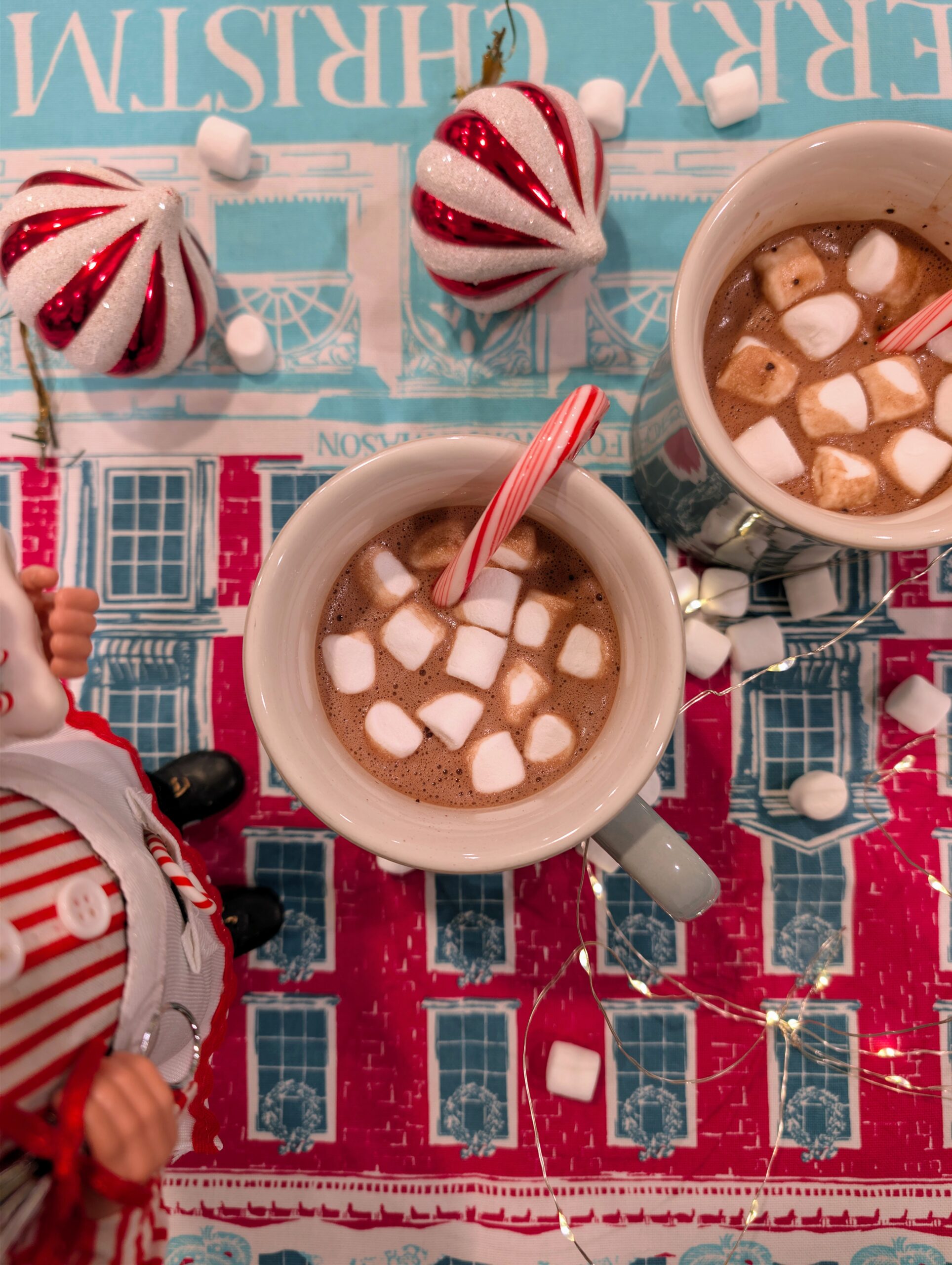 Overhead view of two mugs of homemade hot chocolate topped with marshmallows and candy canes on a festive red and blue holiday table.