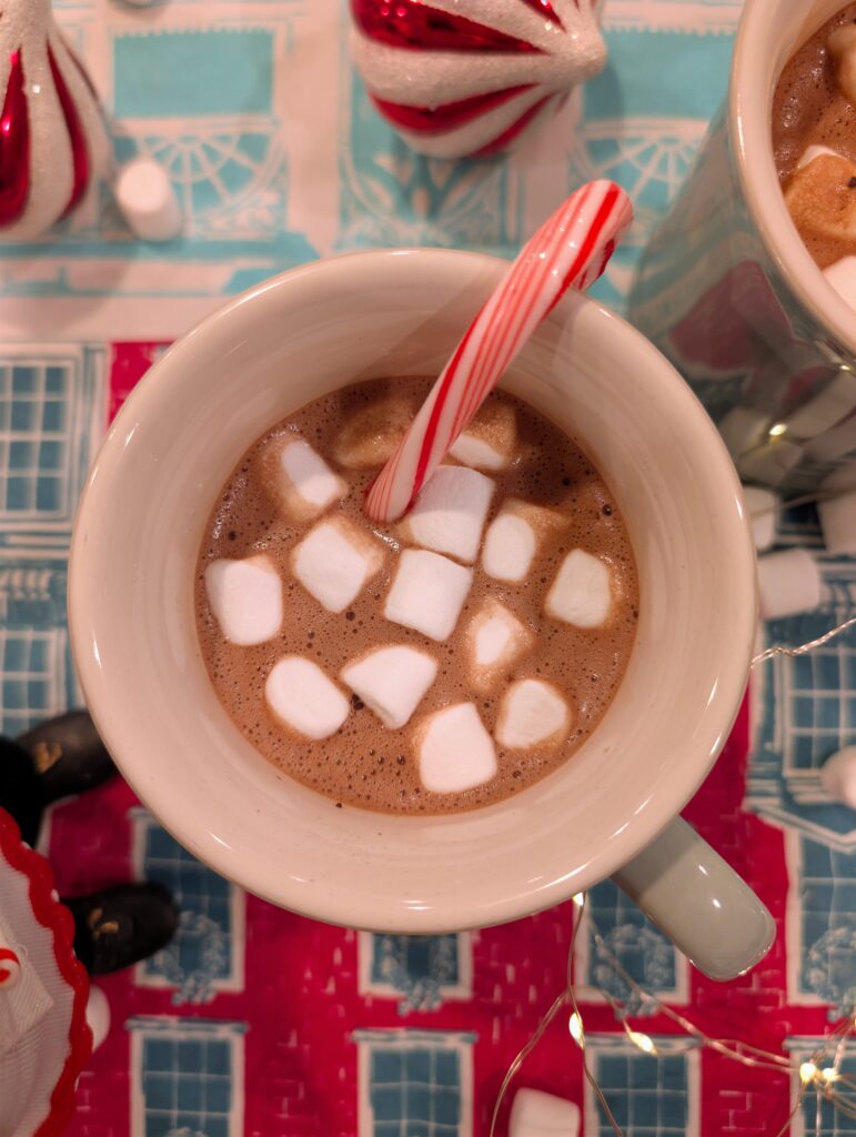 Overhead view of two mugs of homemade hot chocolate topped with marshmallows and candy canes on a festive red and blue holiday table.
