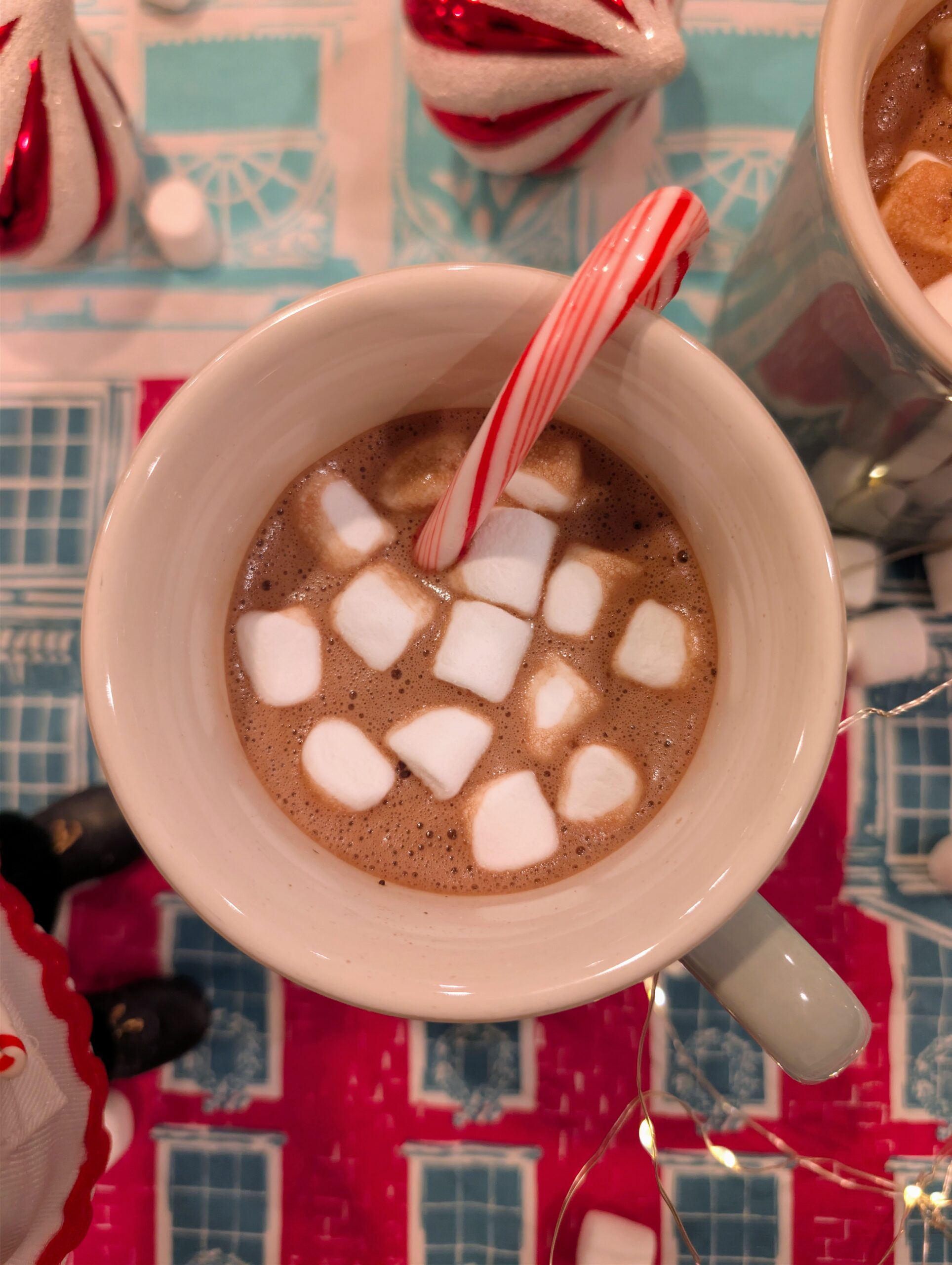 A white ceramic mug filled with hot cocoa and marshmallows sitting next to Christmas ornaments and fairy lights.