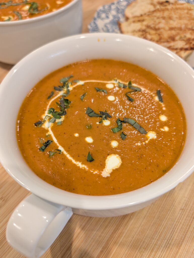 Creamy tomato soup served in a white mug with a side of toasted bread on a wooden surface.
