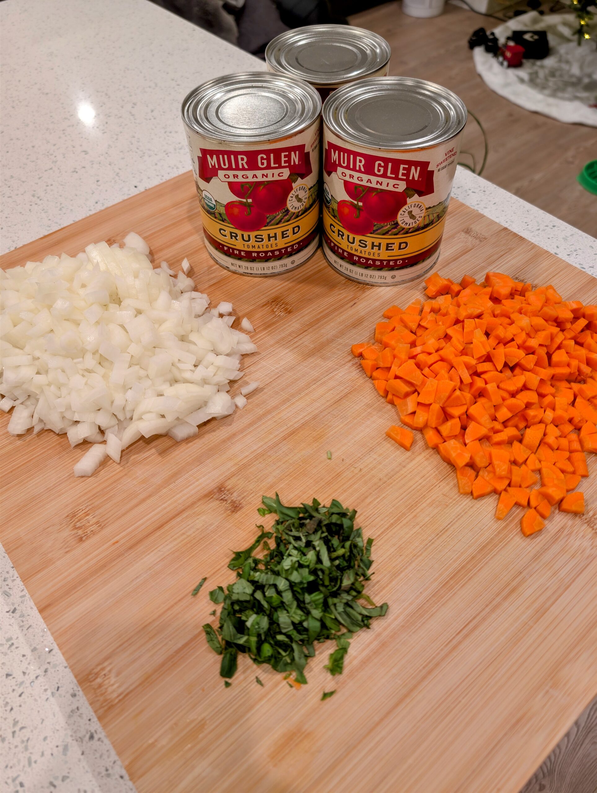 Diced onions, chopped carrots, and fresh herbs on a wooden cutting board next to two cans of crushed tomatoes.