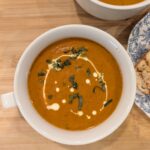 Overhead view of creamy tomato soup in a white bowl garnished with cream and herbs.