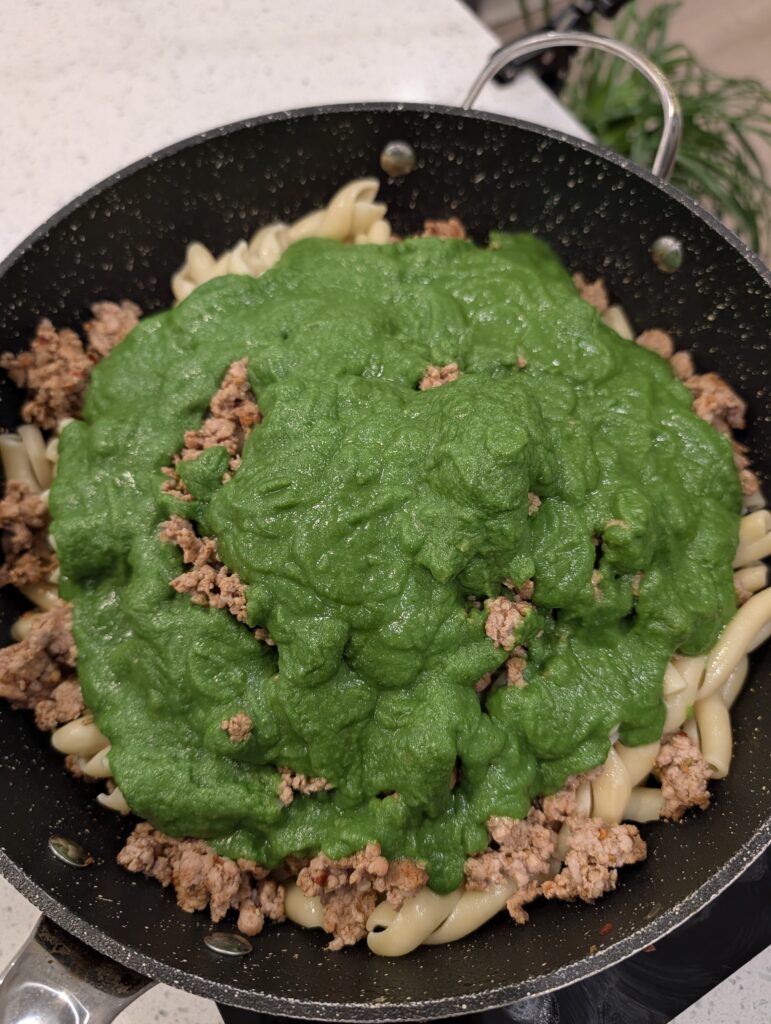 Bright green goddess sauce poured over cooked pasta and Italian sausage in a large black skillet.