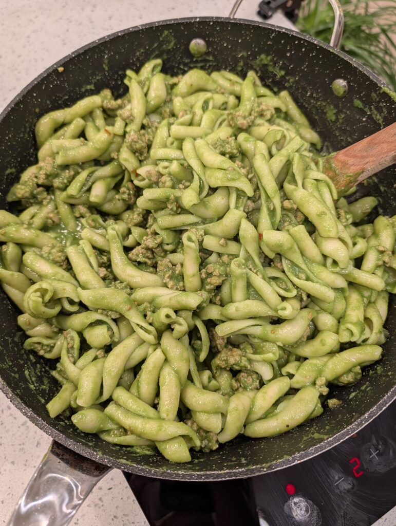 Pasta and crumbled sausage tossed together with a vibrant green vegetable sauce in a pan.