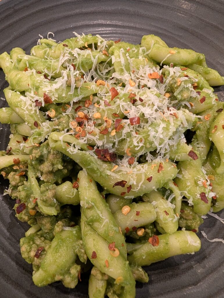 Close up of pasta coated in green sauce and topped with grated parmesan cheese and red pepper flakes.