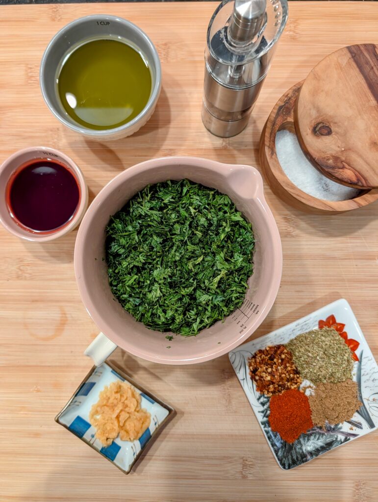 Overhead flat lay of carrot top chimichurri in a pink bowl surrounded by spices, minced garlic, and red wine vinegar on a wooden board