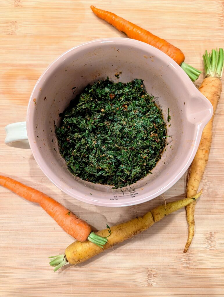 Overhead view of carrot top chimichurri in a pink ceramic bowl with rainbow carrots on a wooden cutting board