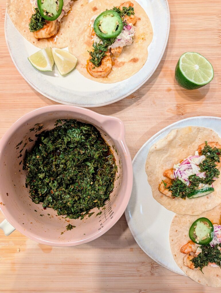 A bowl of carrot top chimichurri on a wooden board alongside shrimp tacos with lime wedges