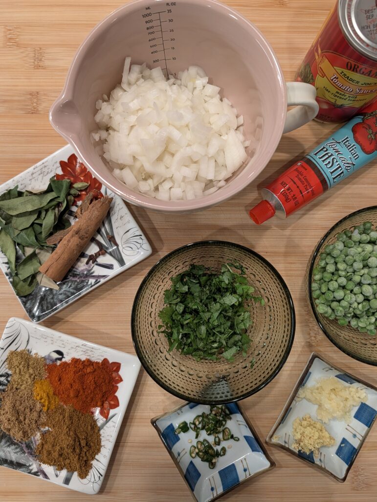 Overhead flat lay of lamb keema ingredients including diced onion, chopped herbs, ground spices, green chili, ginger, garlic, tomato paste, and whole spices on a wooden cutting board