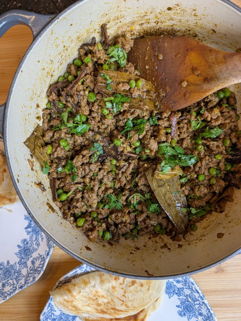 Overhead view of finished lamb keema matar with green peas and fresh cilantro in a large grey Dutch oven, with naan bread on a blue and white plate alongside