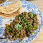 Close-up overhead shot of lamb keema matar topped with fresh cilantro and mint on a blue and white floral plate with paratha on the side