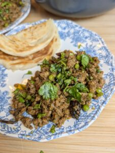 Close-up overhead shot of lamb keema matar topped with fresh cilantro and mint on a blue and white floral plate with paratha on the side