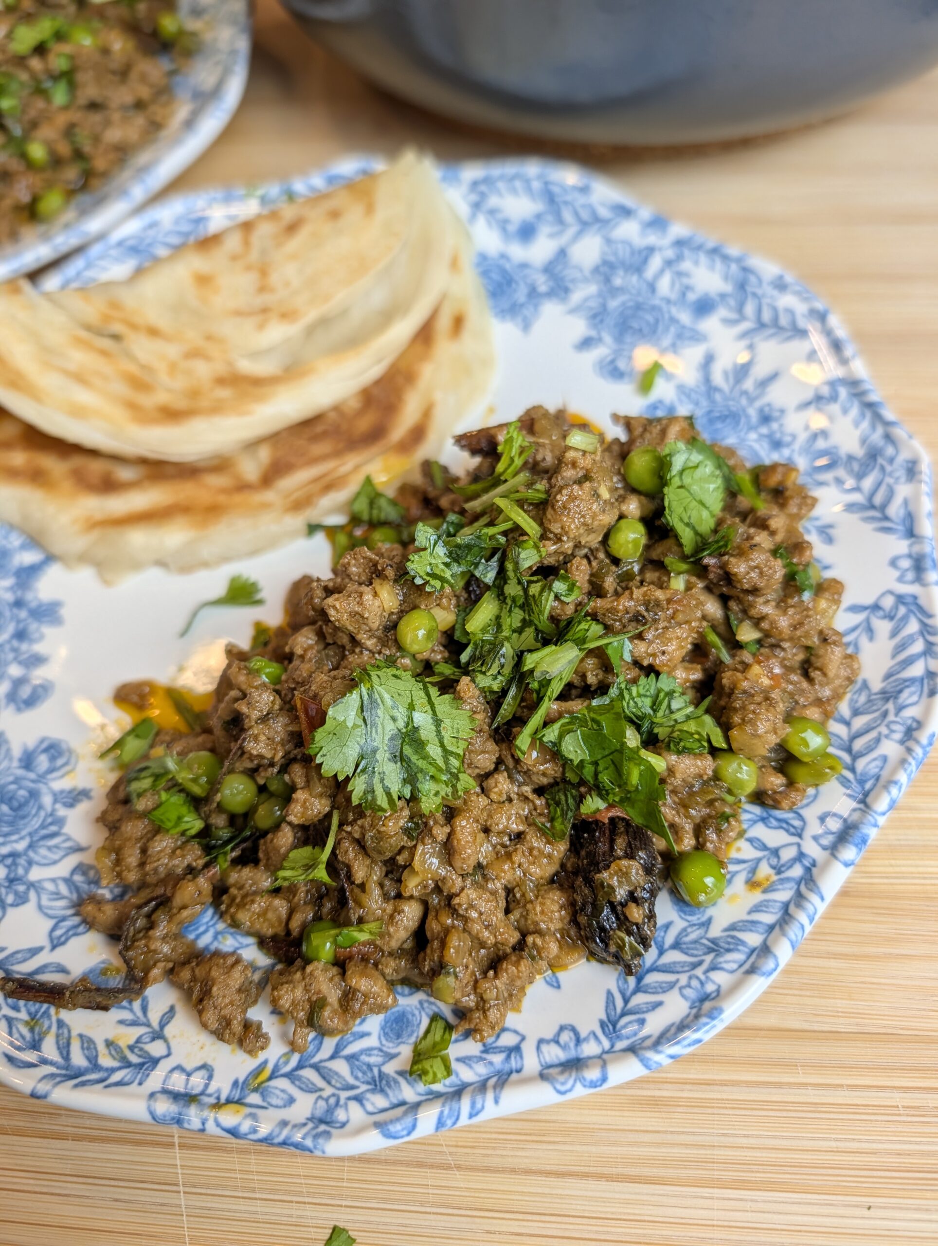Close-up overhead shot of lamb keema matar topped with fresh cilantro and mint on a blue and white floral plate with paratha on the side