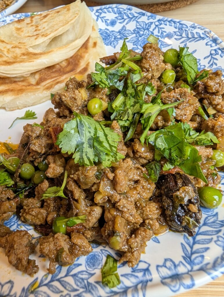 Close-up overhead shot of lamb keema matar topped with fresh cilantro and mint on a blue and white floral plate with paratha on the side