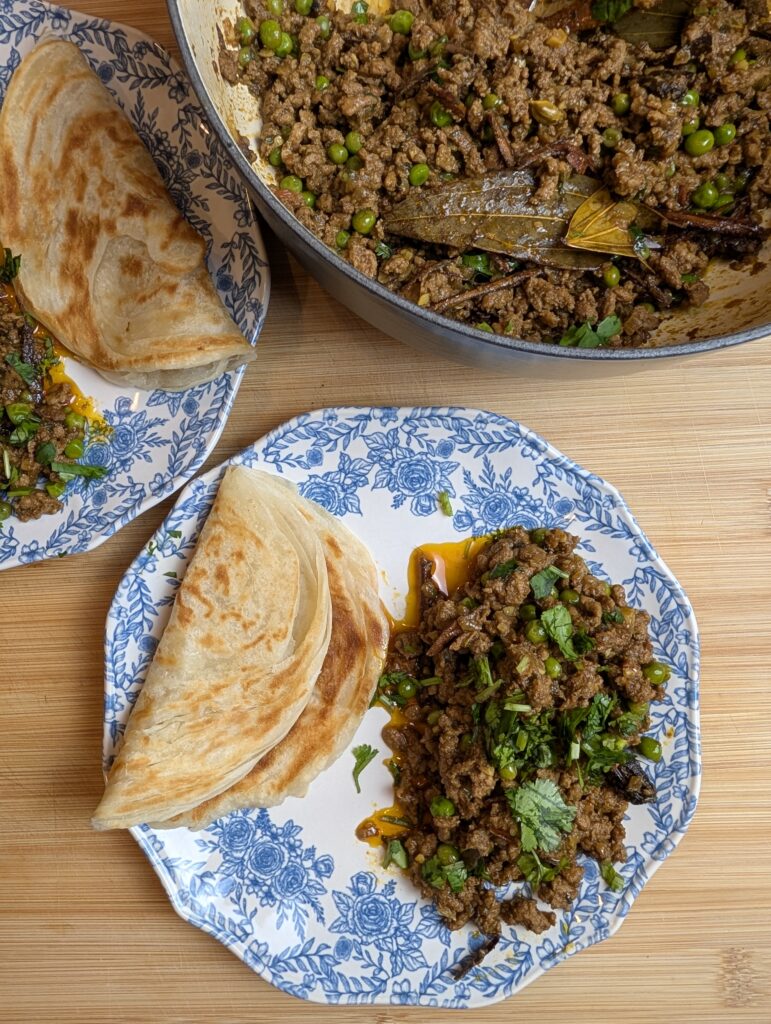 Overhead shot of a blue and white plate with lamb keema matar and flaky paratha alongside a large pot of keema on a wooden surface