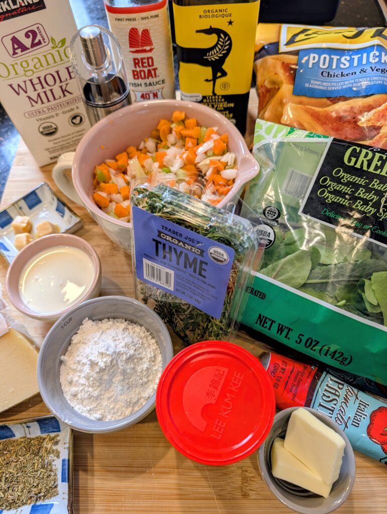 Overhead flat lay of chicken dumpling soup ingredients including diced mirepoix vegetables, flour, cream, fresh thyme, spinach, tomato paste, frozen potstickers, and chicken broth on a wooden surfac
