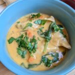 Close-up overhead shot of creamy chicken dumpling soup in a blue bowl topped with fresh parsley, with dumplings and wilted spinach visible in a golden broth