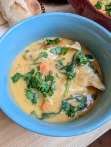 Close-up overhead shot of creamy chicken dumpling soup in a blue bowl topped with fresh parsley, with dumplings and wilted spinach visible in a golden broth