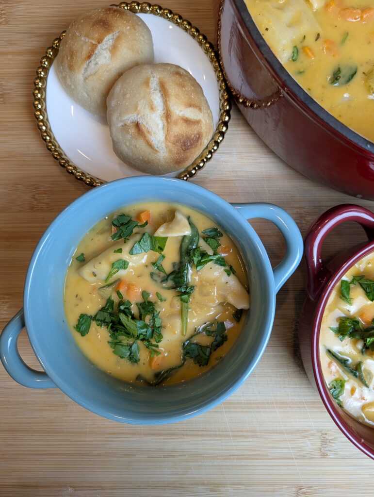 Overhead shot of creamy chicken dumpling soup in a blue bowl alongside a small red pot of soup, a large red Dutch oven, and crusty rolls on a gold-rimmed plate on a wooden surface