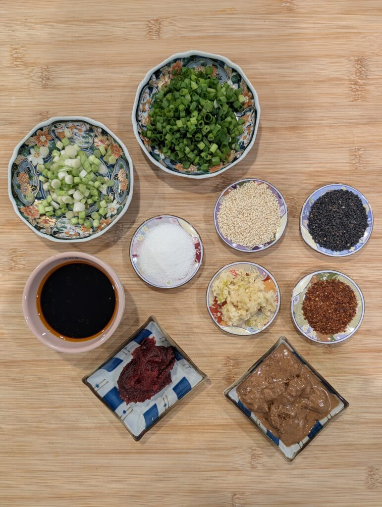 Overhead flat lay of gochujang peanut butter noodle ingredients including scallion whites and greens, white and black sesame seeds, sugar, soy sauce, minced garlic, gochugaru, gochujang, and peanut butter arranged in small bowls on a wooden surface