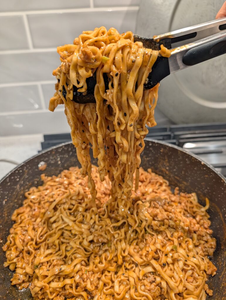 Action shot of gochujang peanut butter noodles with ground pork being tossed with tongs in a dark skillet on a gas stovetop