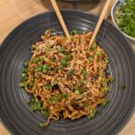 Overhead shot of two dark bowls of gochujang peanut butter noodles topped with scallions and sesame seeds with chopsticks, alongside a small bowl of scallion greens on a wooden surface