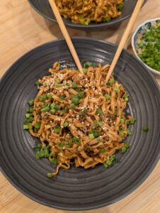 Overhead shot of two dark bowls of gochujang peanut butter noodles topped with scallions and sesame seeds with chopsticks, alongside a small bowl of scallion greens on a wooden surface