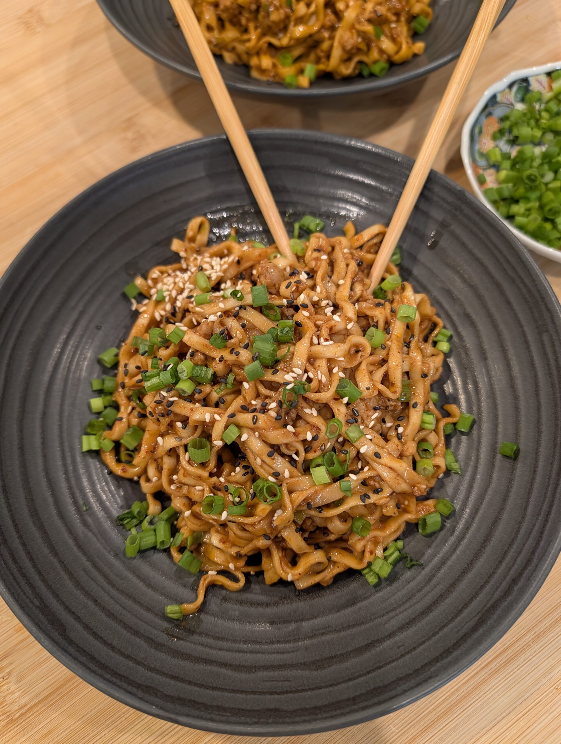 Overhead shot of two dark bowls of gochujang peanut butter noodles topped with scallions and sesame seeds with chopsticks, alongside a small bowl of scallion greens on a wooden surface