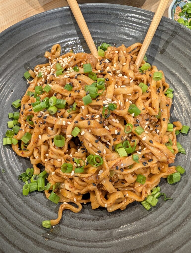 Overhead close-up of gochujang peanut butter noodles in a dark bowl topped with scallion greens and white and black sesame seeds with chopsticks resting on top
