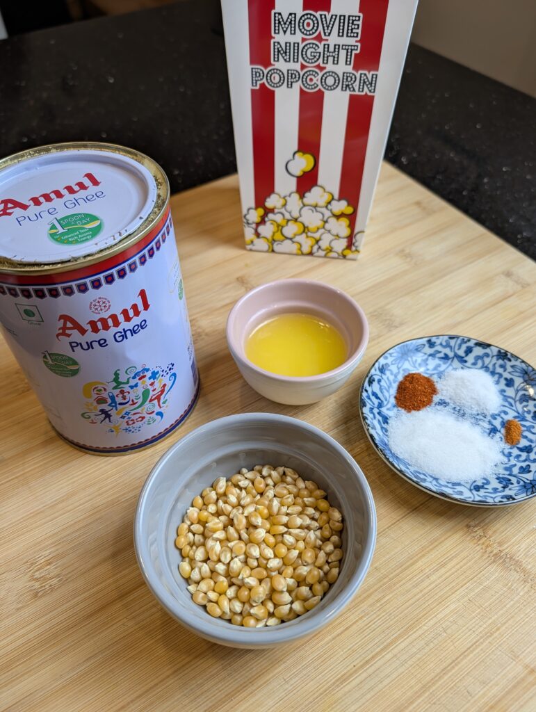 Overhead flat lay of sweet and salty popcorn ingredients including popcorn kernels, melted ghee, sugar, salt, paprika, and cayenne alongside a movie night popcorn box on a wooden surface