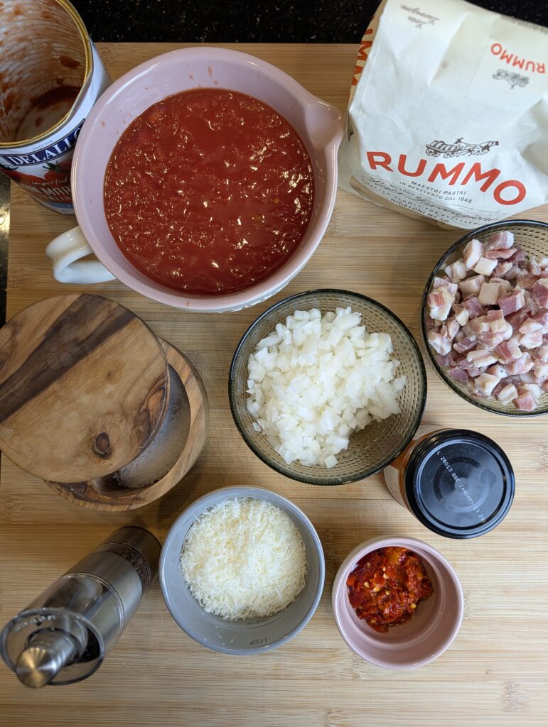 Ingredients for amatriciana pasta including guanciale, diced onion, tomato sauce, grated cheese, and Rummo pasta on a counter.