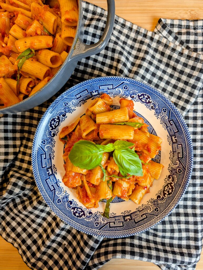 Looking down at a portion of amatriciana pasta garnished with fresh basil on a patterned plate over a plaid napkin.