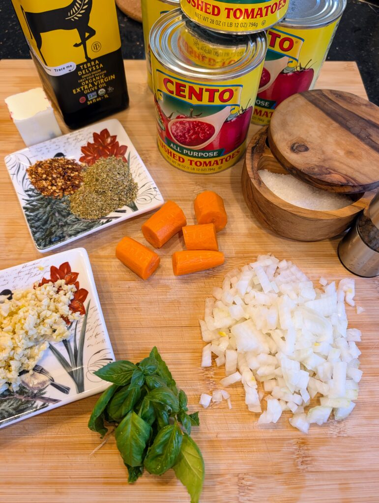 Overhead flat lay of red sauce ingredients including cans of crushed tomatoes, olive oil, butter, diced onion, carrot chunks, minced garlic, fresh basil, red pepper flakes, and dried oregano on a wooden cutting board