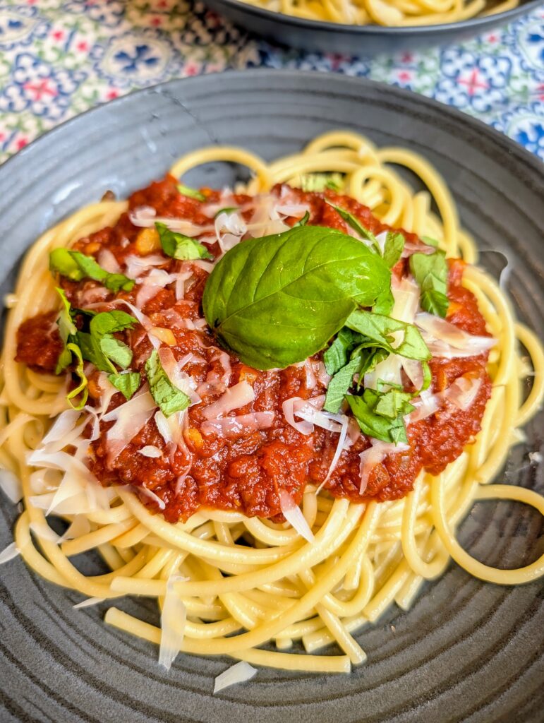 Overhead shot of spaghetti topped with slow-simmered red sauce, shaved parmesan, and fresh basil on a dark plate with a second plate visible in the background on a blue patterned tablecloth