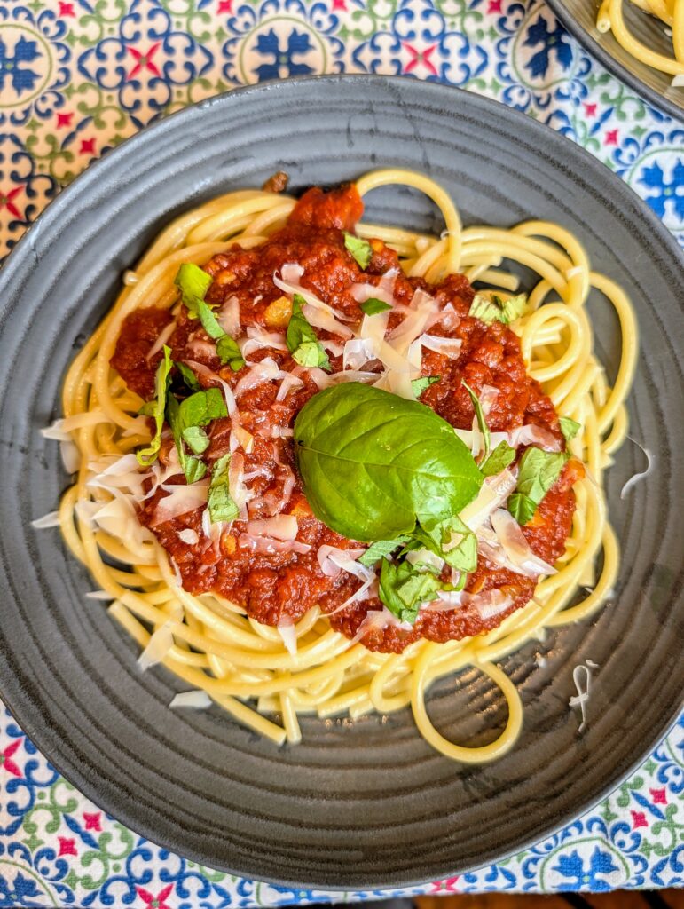 Overhead shot of spaghetti topped with slow-simmered red sauce, shaved parmesan, and fresh basil on a dark plate with a second plate visible in the background on a blue patterned tablecloth