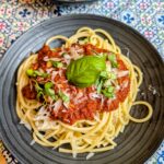 Overhead shot of spaghetti topped with slow-simmered red sauce, shaved parmesan, and fresh basil on a dark plate with a second plate visible in the background on a blue patterned tablecloth