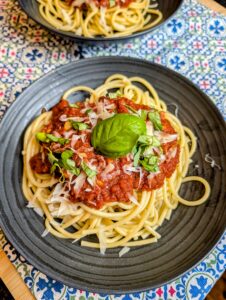 Overhead shot of spaghetti topped with slow-simmered red sauce, shaved parmesan, and fresh basil on a dark plate with a second plate visible in the background on a blue patterned tablecloth