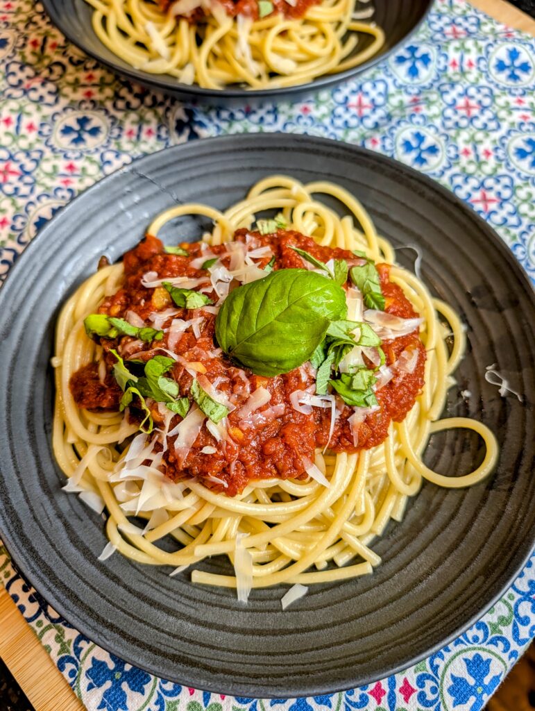 Overhead shot of spaghetti topped with slow-simmered red sauce, shaved parmesan, and fresh basil on a dark plate with a second plate visible in the background on a blue patterned tablecloth