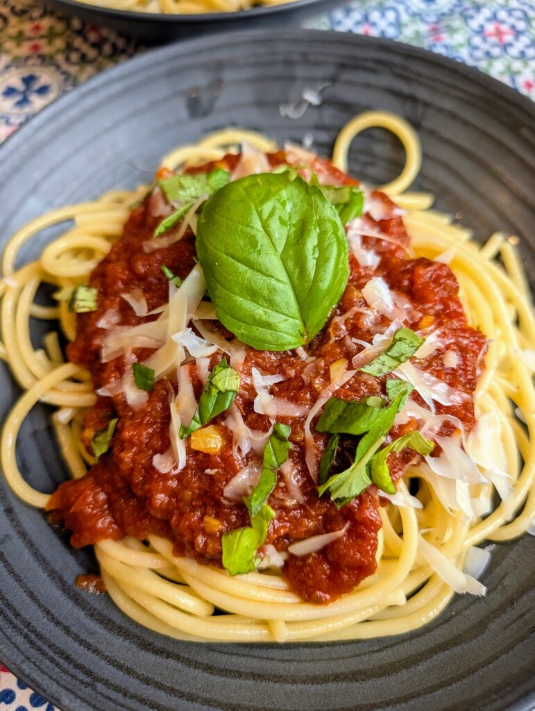 Close-up overhead shot of spaghetti with rich red tomato sauce topped with a whole basil leaf, shaved parmesan, and fresh chopped basil on a dark plate